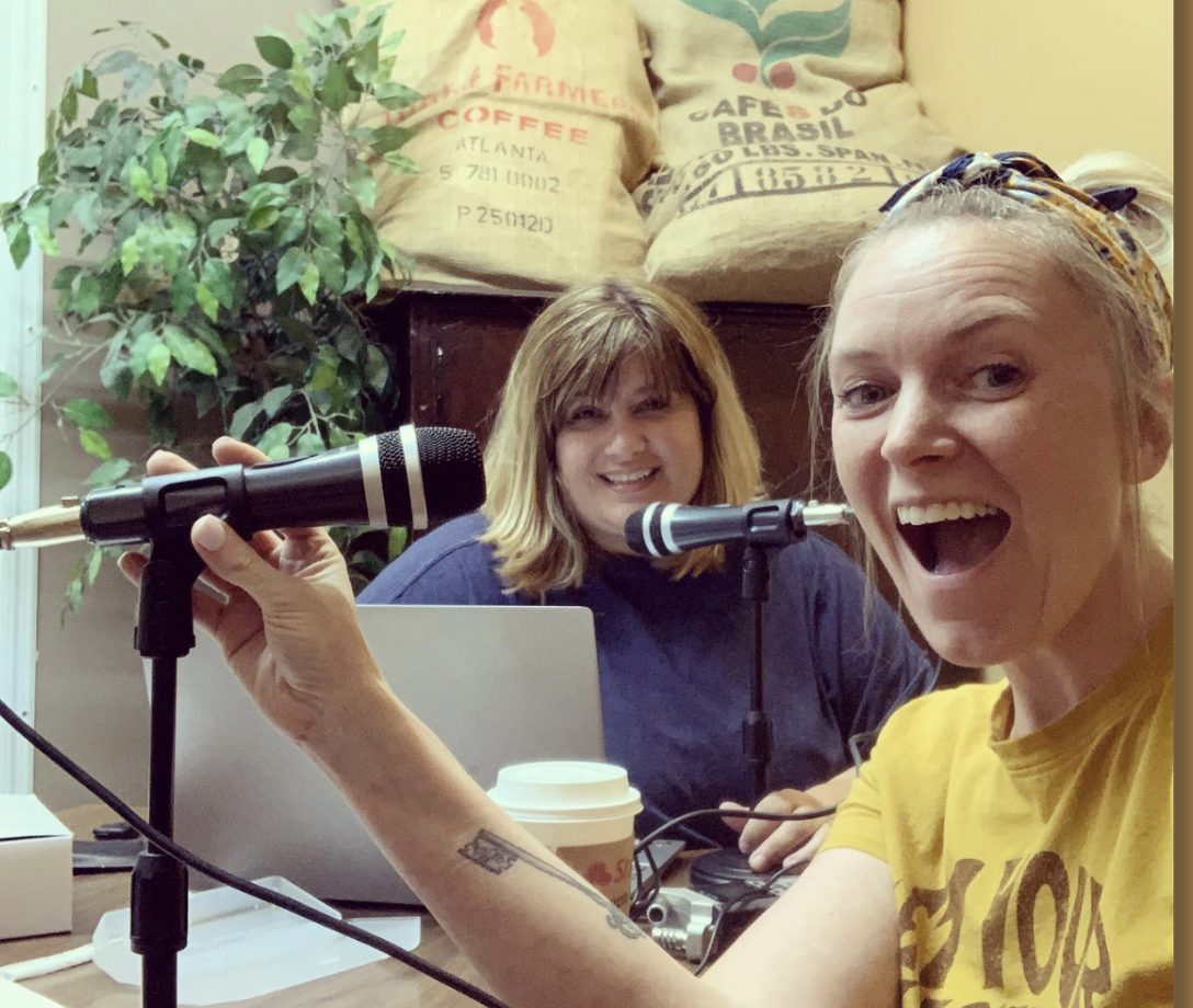 Amarilys and Misty at their first recording session in 2020, laughing behind two microphones, a laptop and a coffee cup, with burlap coffee sacks on the wall behind them.